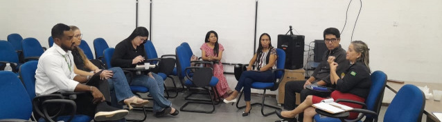 Pessoas participando de uma reunião em uma sala, sentadas em cadeiras azuis dispostas em círculo, discutindo um tema.