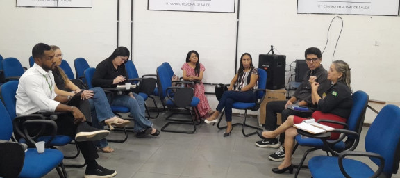 Pessoas participando de uma reunião em uma sala, sentadas em cadeiras azuis dispostas em círculo, discutindo um tema.