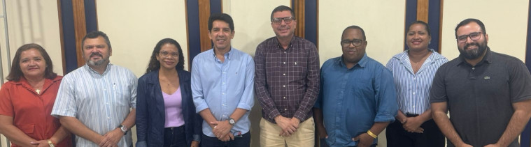 Oito pessoas estão lado a lado, posando para uma foto em pé. Há homens e mulheres, de diferentes tons de pele, usando roupas variadas. Eles estão em uma sala com paredes claras e detalhes em madeira. Todos sorriem para a câmera.
