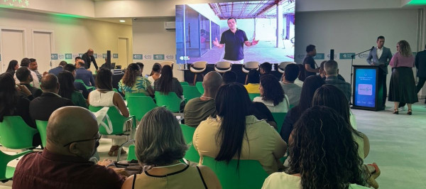Sala de evento com participantes sentados em cadeiras verdes, assistindo a uma apresentação em um telão iluminado.